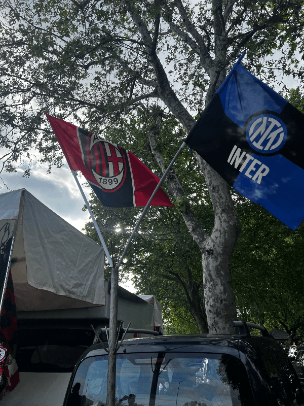 AC Milan and Inter Milan rival club flags displayed together outdoors, showing the AC Milan crest with the 1899 founding year alongside the blue and black Inter Milan flag