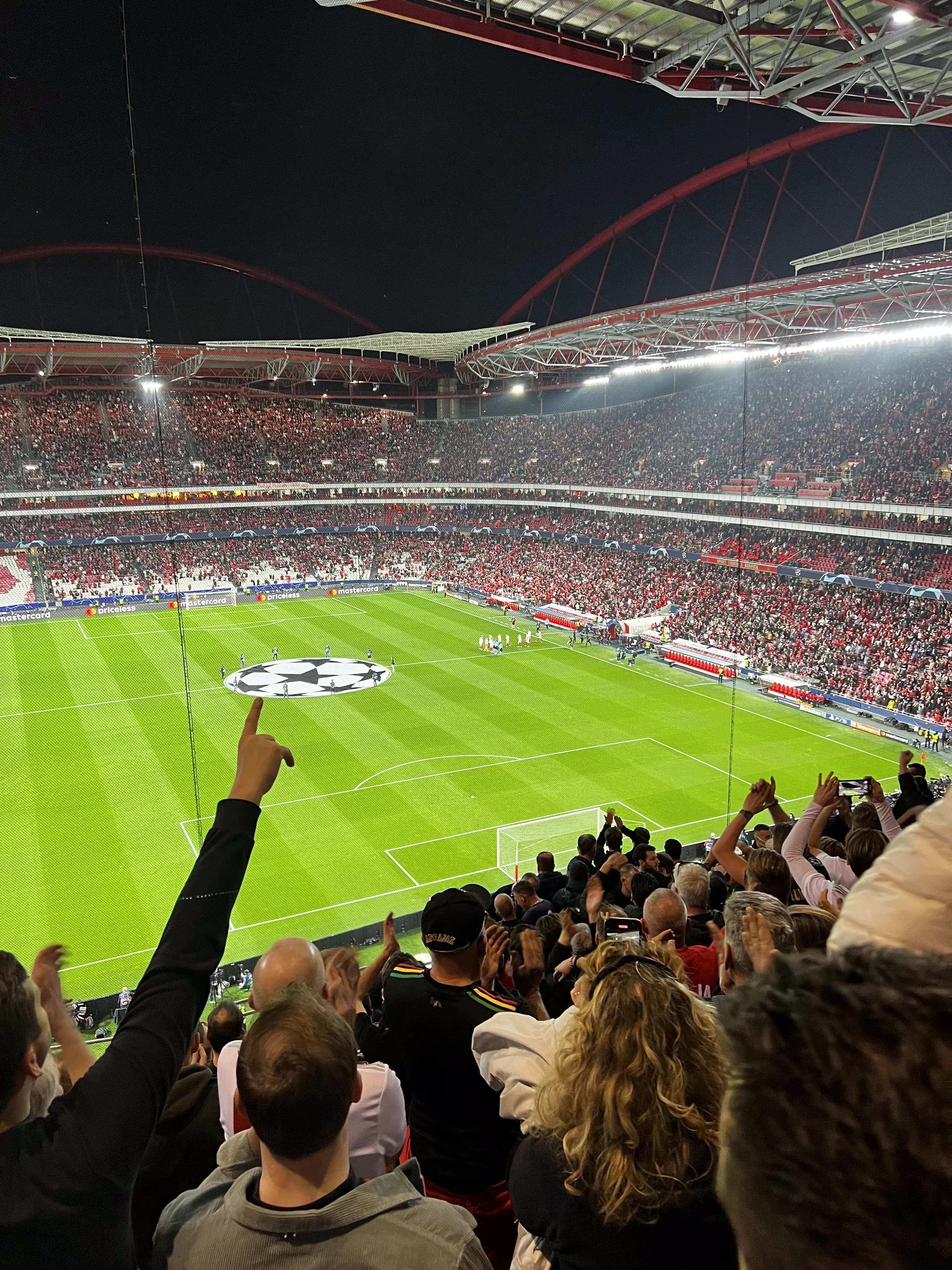 Away fans cheering inside a packed Estádio da Luz during a UEFA Champions League night match, with the pitch and center circle logo visible below.