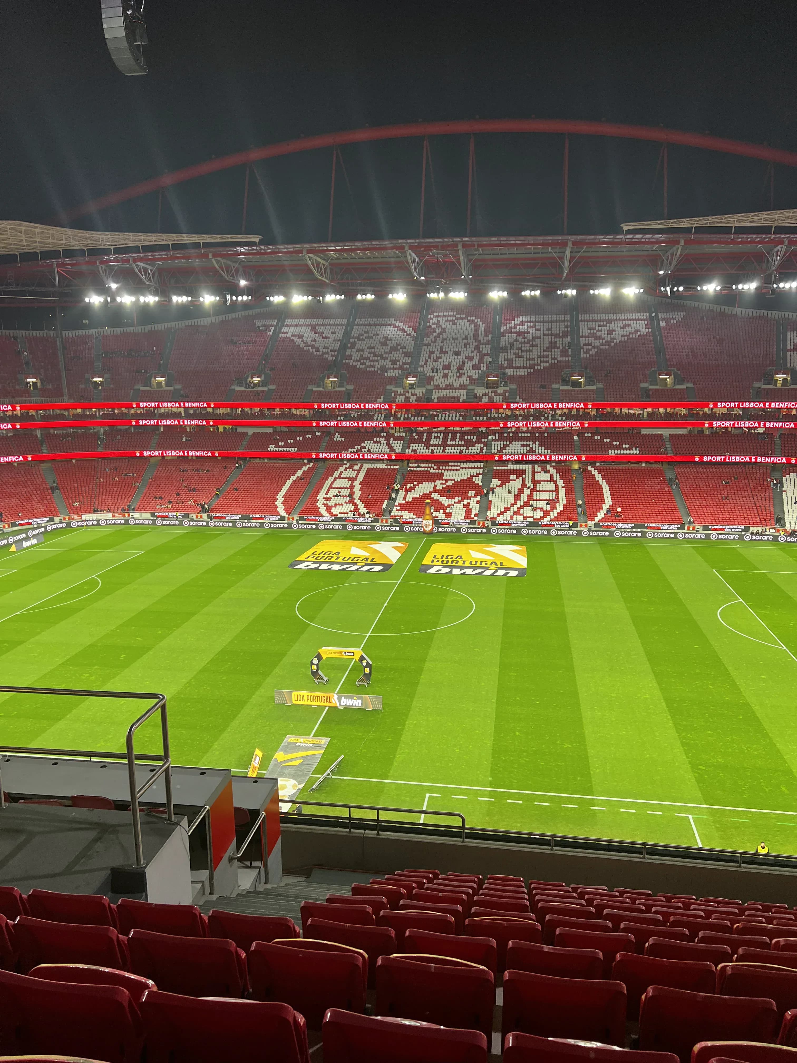 Empty interior of Estádio da Luz at night showing the full pitch, red seating, and the Benfica eagle mosaic in the stands before a Liga Portugal match