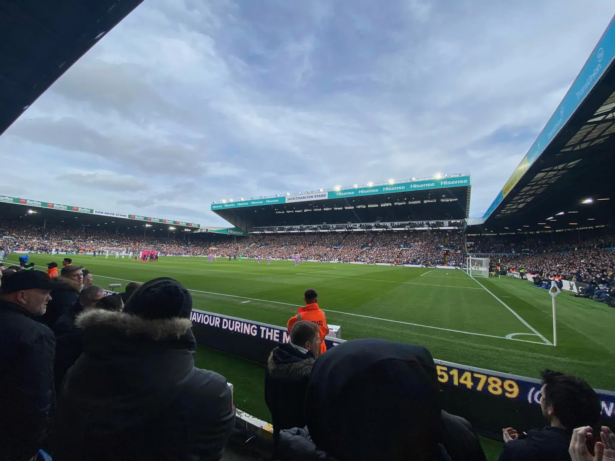 Corner view from the lower tier at Elland Road during a match with a packed Hisense-branded stand in the background under moody skies