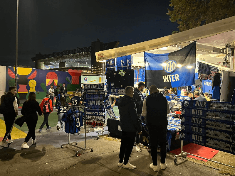 Inter Milan merchandise stall outside San Siro stadium at night, displaying blue and black scarves, jerseys including Crespo number 9, and Inter Milan flags before a matchday
