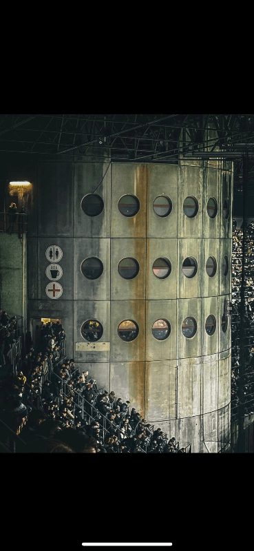 Inter Milan fans packed into San Siro stadium stands at night, with the iconic circular porthole windows of the stadium's brutalist concrete interior visible alongside restroom, café, and first aid signs